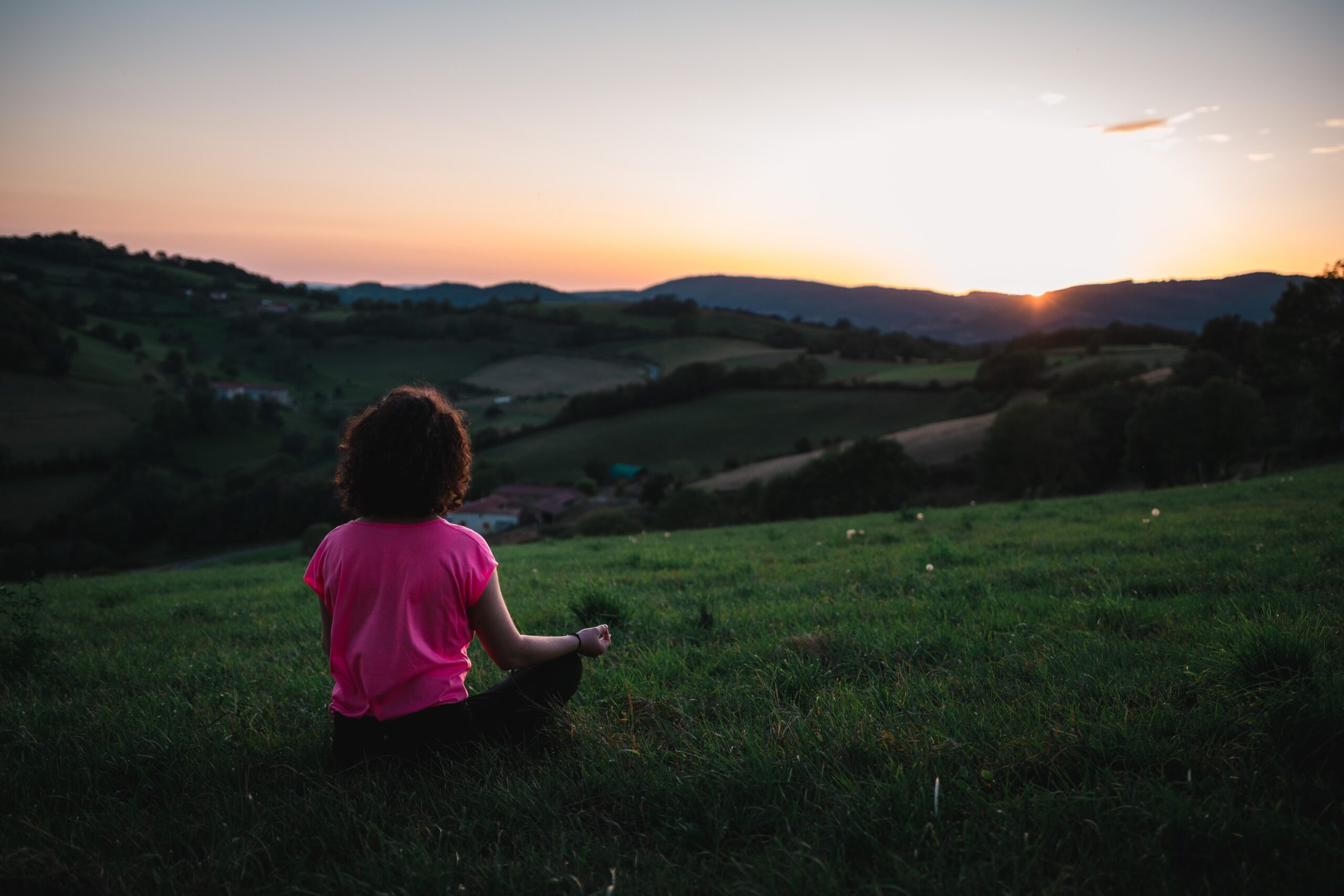 light brown skinned woman with dark hair in pink t-shirt sitting in meditation position in brightly lit green field overlooking sunrise and mountains in the distance