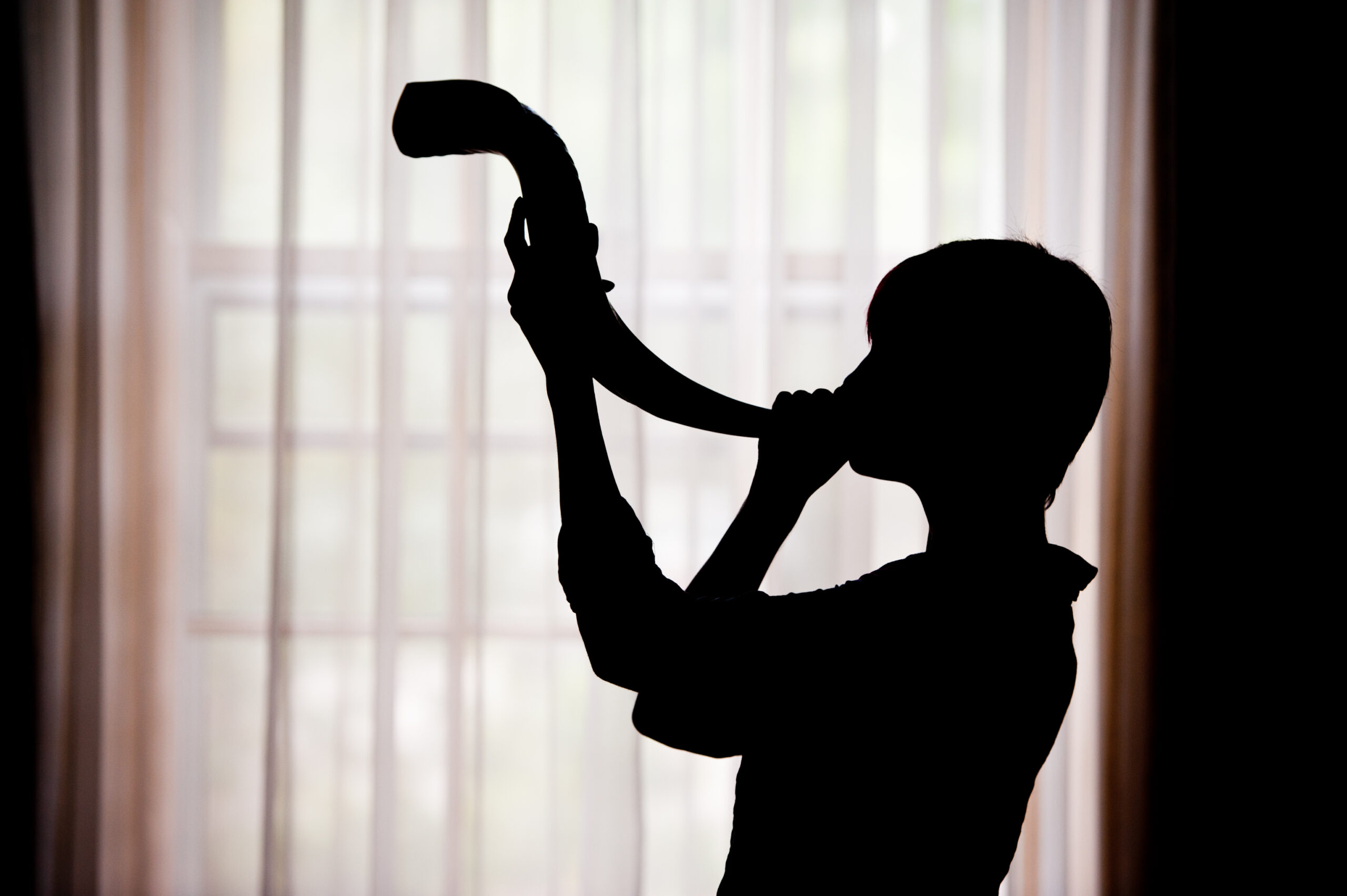 person in silhouette blowing shofar against white curtains