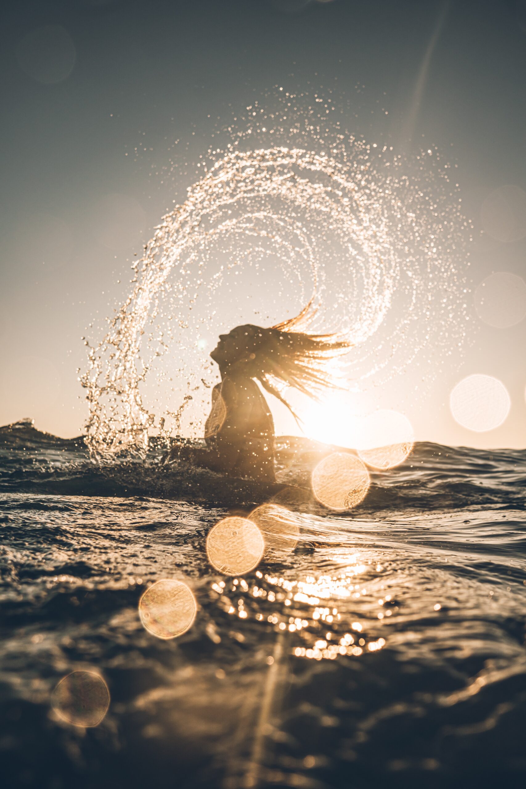 person with long hair emerging from the water splashing an arc of water from their hair against the sunset
