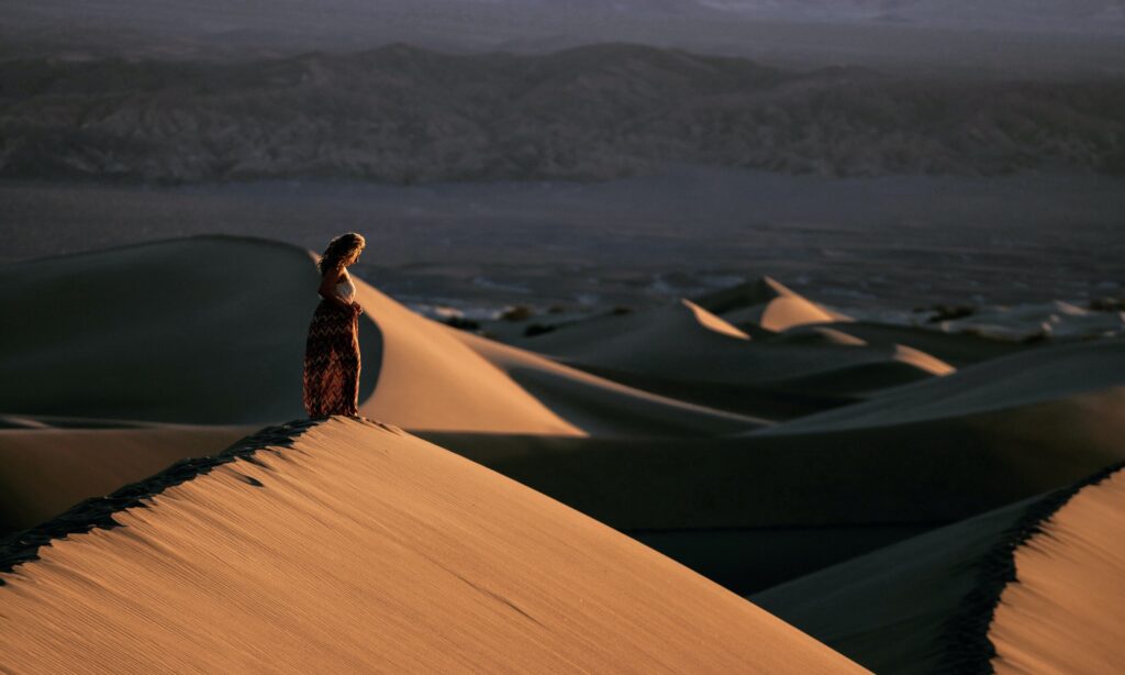 woman in a desert with mounds of sand shown with shadows. she is small and far away in the scene.