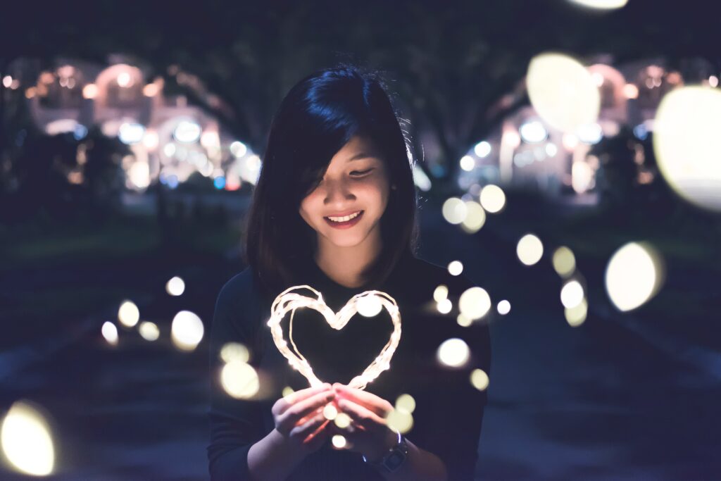 smiling asian kid with black long hair holding a glowing heart that appears to be made out of wire with glowing lights in the background