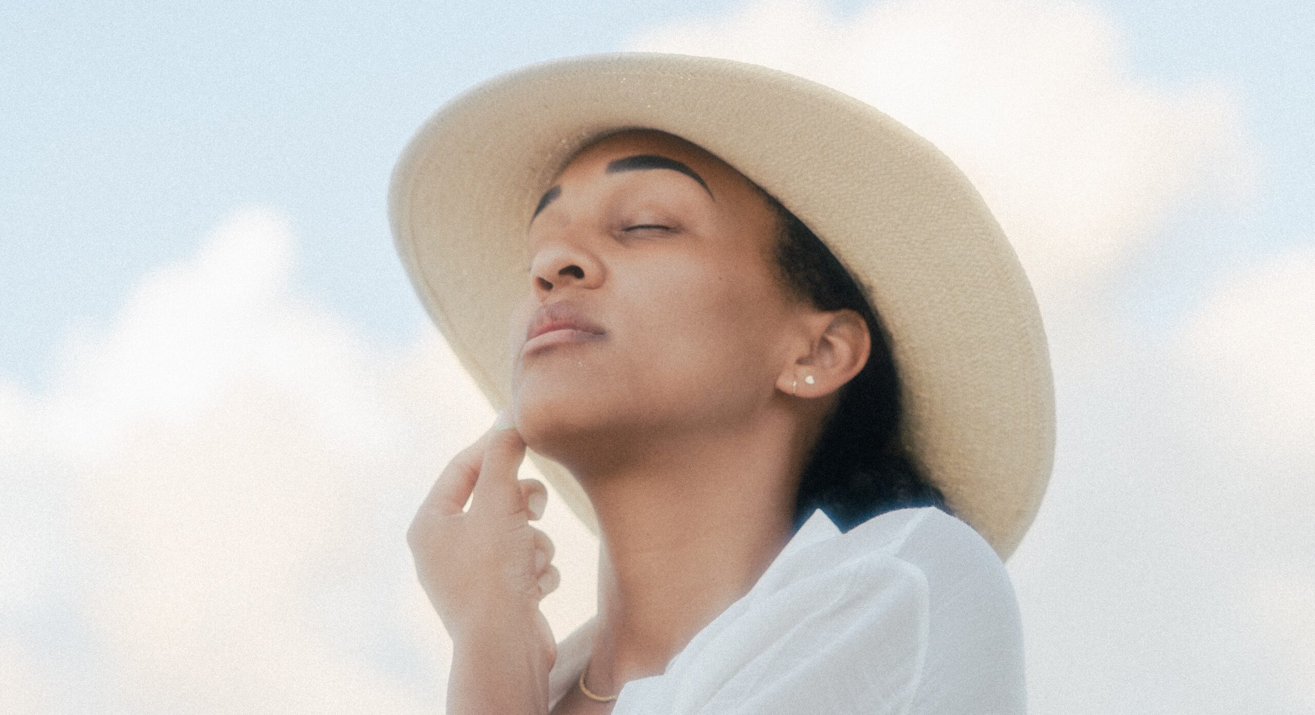 light brown skinned person with eyes closed and white shirt on and beige sun hat with head slightly lifted looking peaceful before light blue sky and clouds