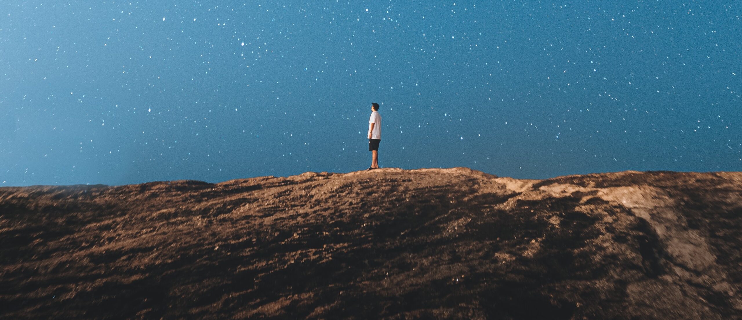 person standing small on rocky land beneath blue sky full of stars