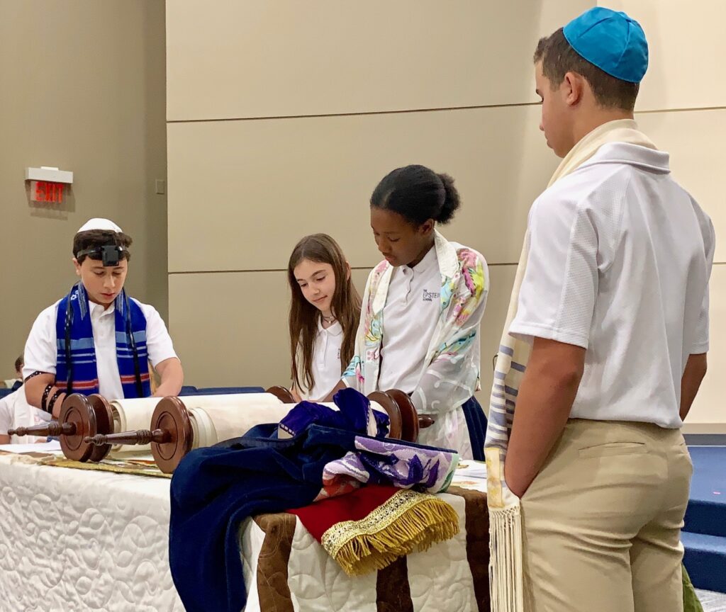 four children standing around an open torah scroll in synagogue. the girl in the center is black and wearing a tallit, looking like she is reading from the torah. one girl looking on is white. Two white boys wearing kippot and tallitot are at the side watching.