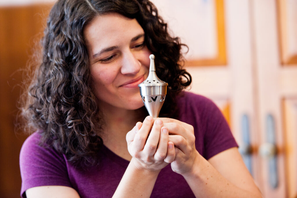 light skinned jewish woman with dark curly hair in purple t-shirt holding a silver spice holder and sniffing the spices with a smile