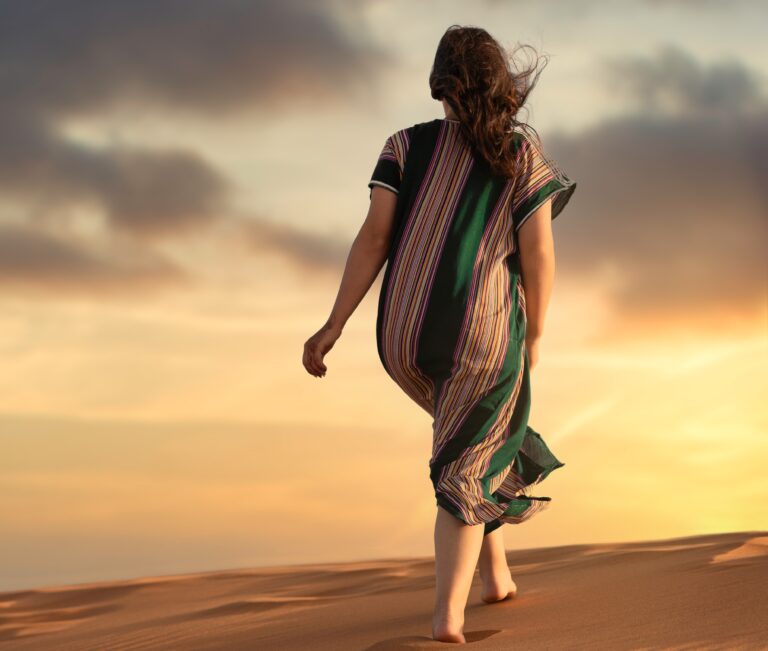 woman walking barefoot in desert wearing loose striped dress with hair blowing in the wind