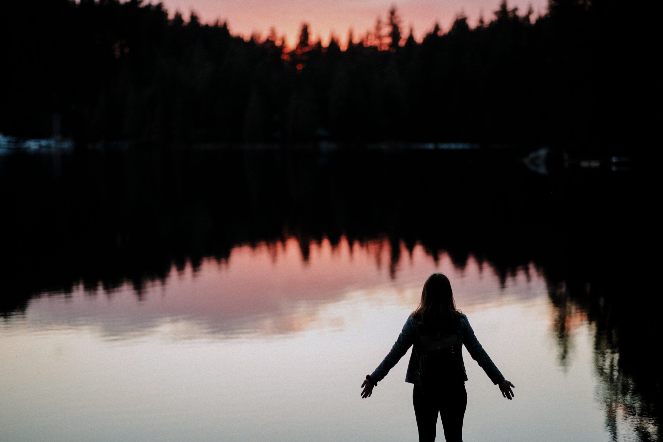 silhouette of woman standing before lake with dark trees in the background reflected in the lake. her arms are spread wide.