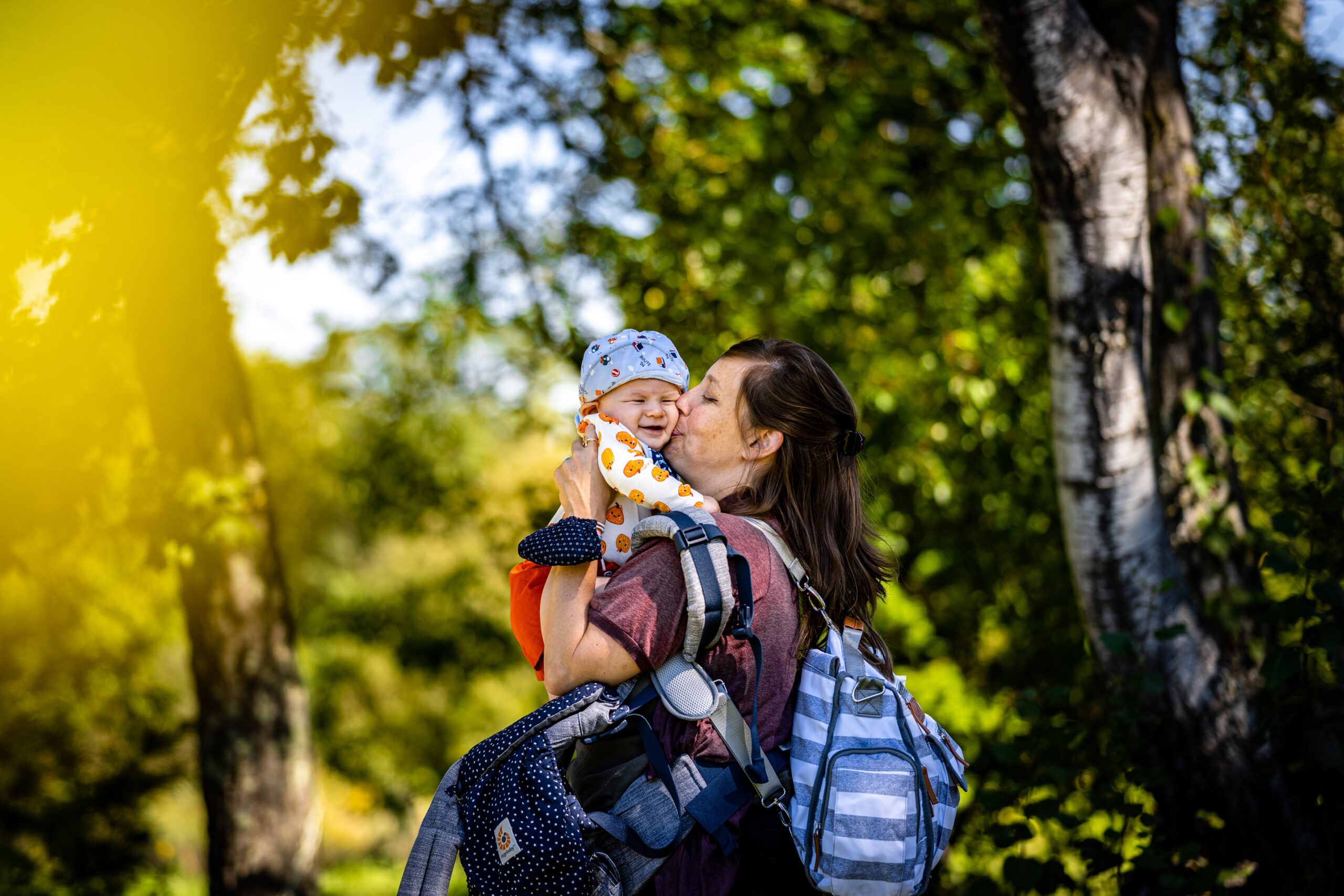 light skinned brown haired mother holding small child and multiple bags and a diaper bag outdoors giving the child a kiss