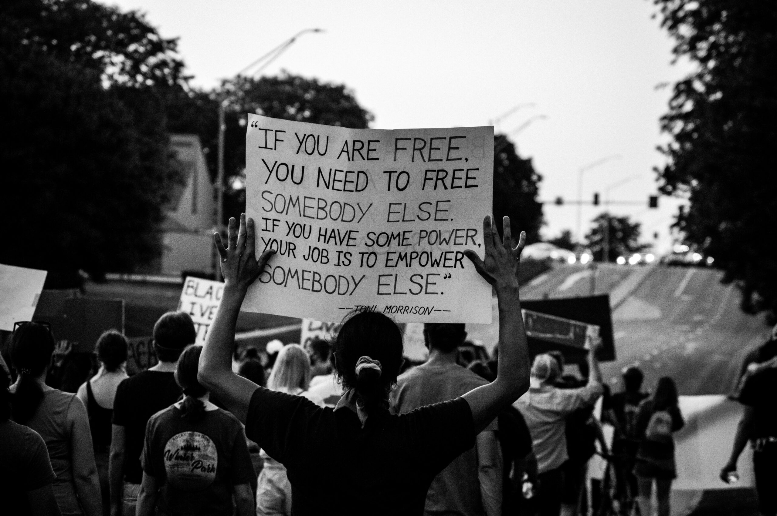 black and white photo of black person at a protest holding up a sign that reads “If you are free, you need to free somebody else. If you have some power, then your job is to empower somebody else.” ―Toni Morrison