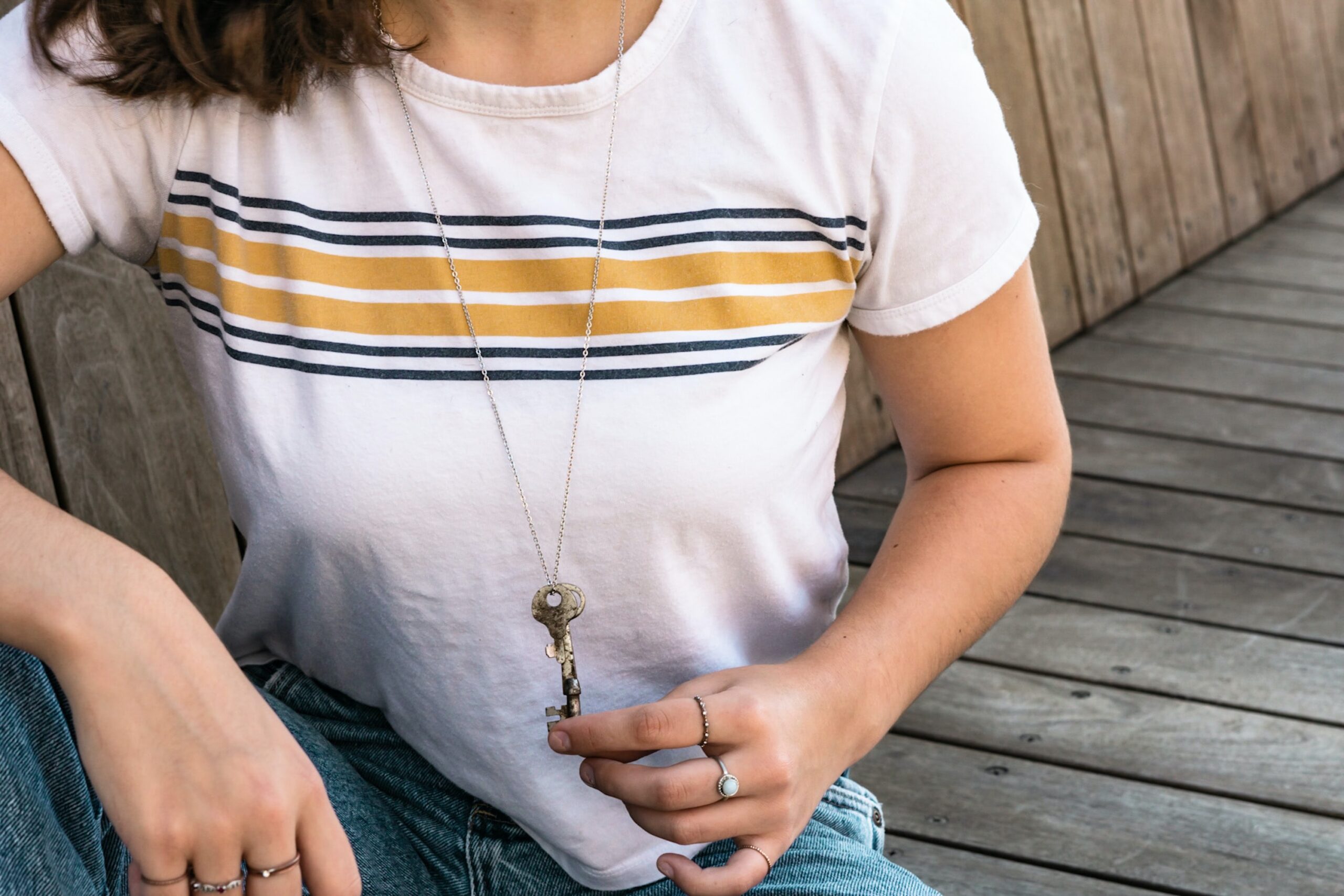 close up of white woman wearing white t-shirt with a few colorful horizontal stripes. she's wearing a necklace with a key hanging on it, and she's holding the key with two fingers. she has rings on her pointer and middle fingers.