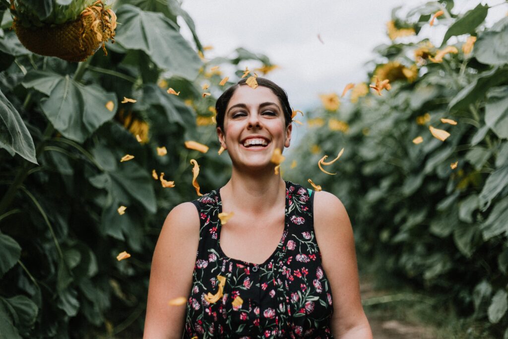 woman with light olive skin and short dark hair in black tank top with colorful pattern smiling, eyes closed, in a field of tall flowers, with yellow petals raining down