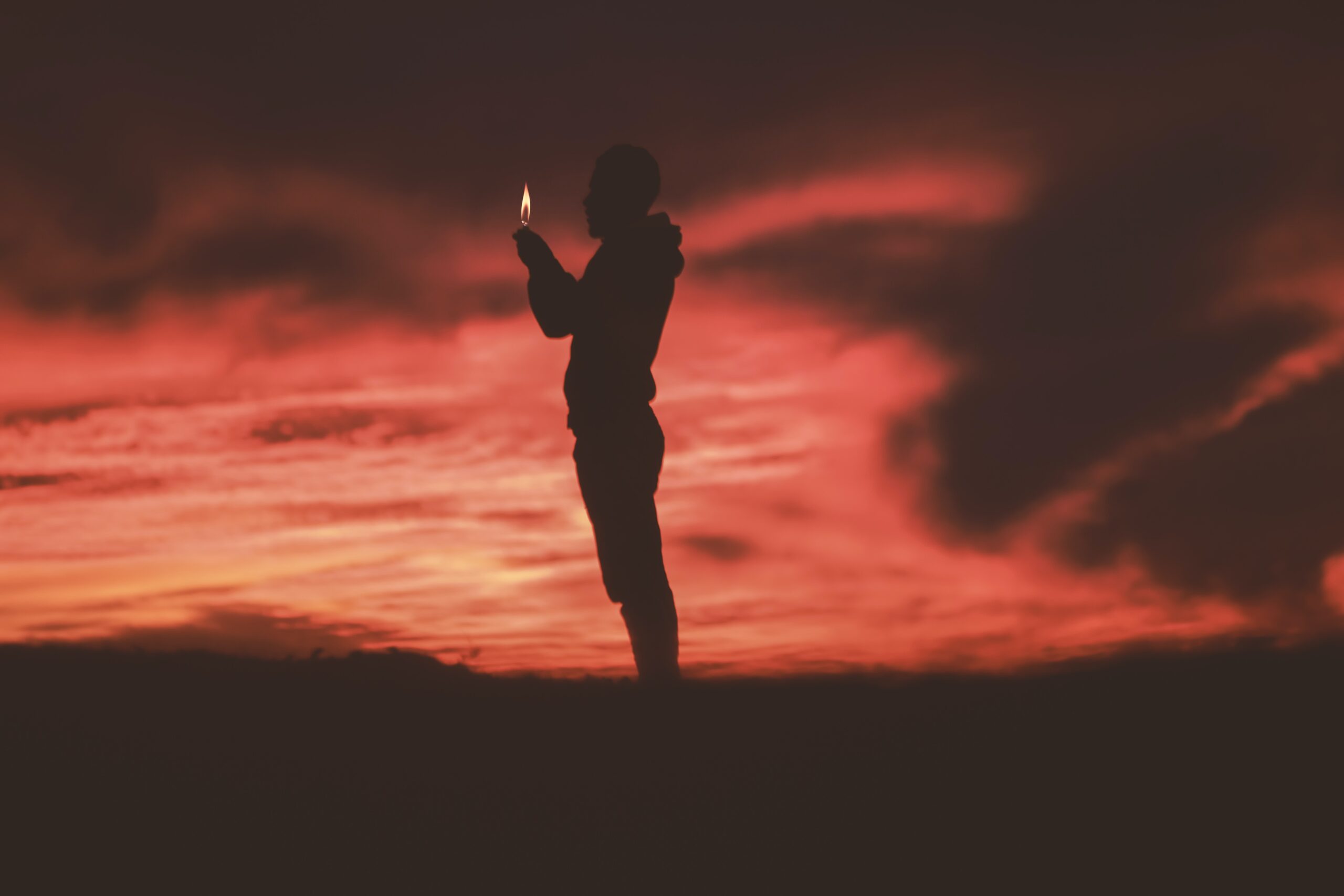 person standing in silhouette against reddish dark sky, holding a flame