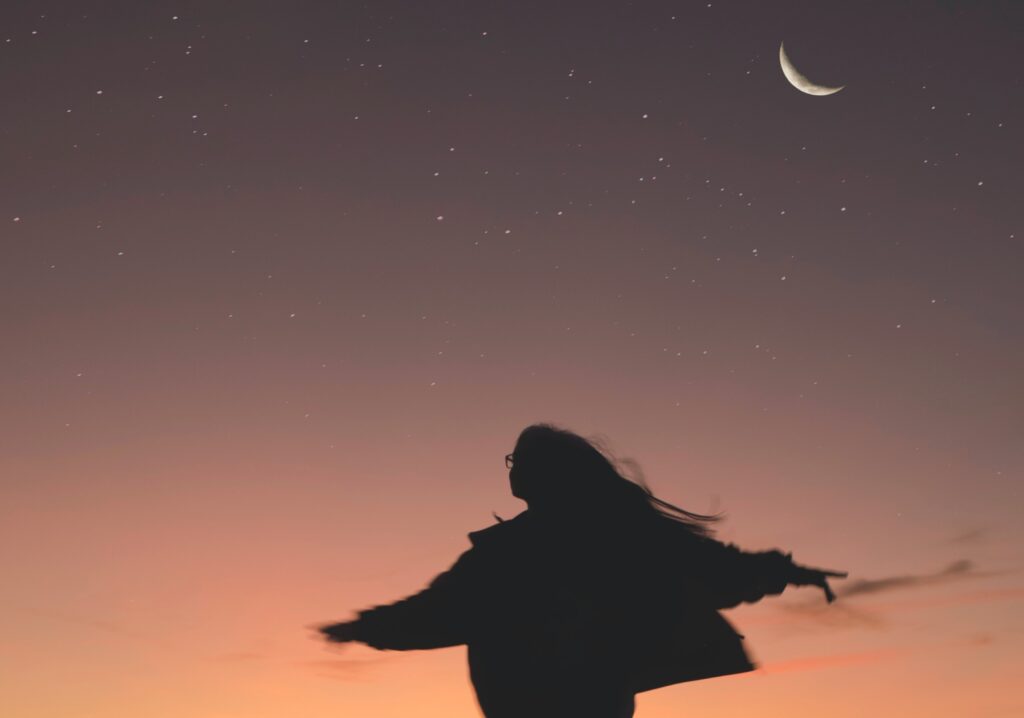 woman in silhouette with arms outspread and hair blowing standing or spinning or dancing in front a purple pink night sky with silver of new moon and stars