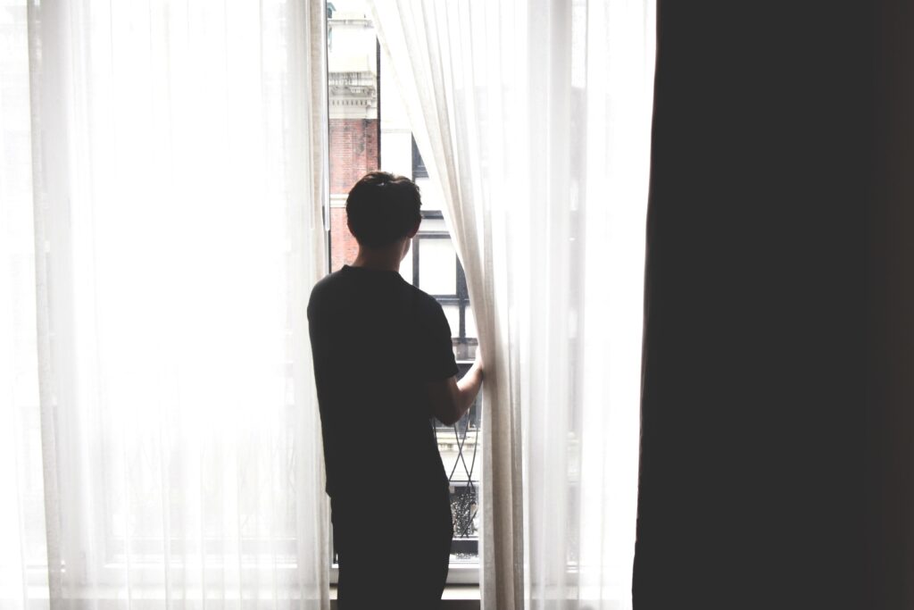 young man in black t-shirt and pants shown from behind standing at curtained window, parting the curtain and looking outside. Curtain is a gauzy white and there is a black area to the right, creating a stark white and black effect to the scene.