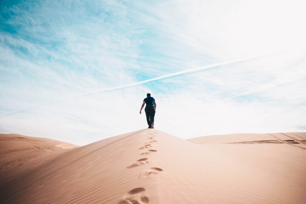 person climbing hill in sandy desert with footsteps behind them and bright blue sky with white clouds above