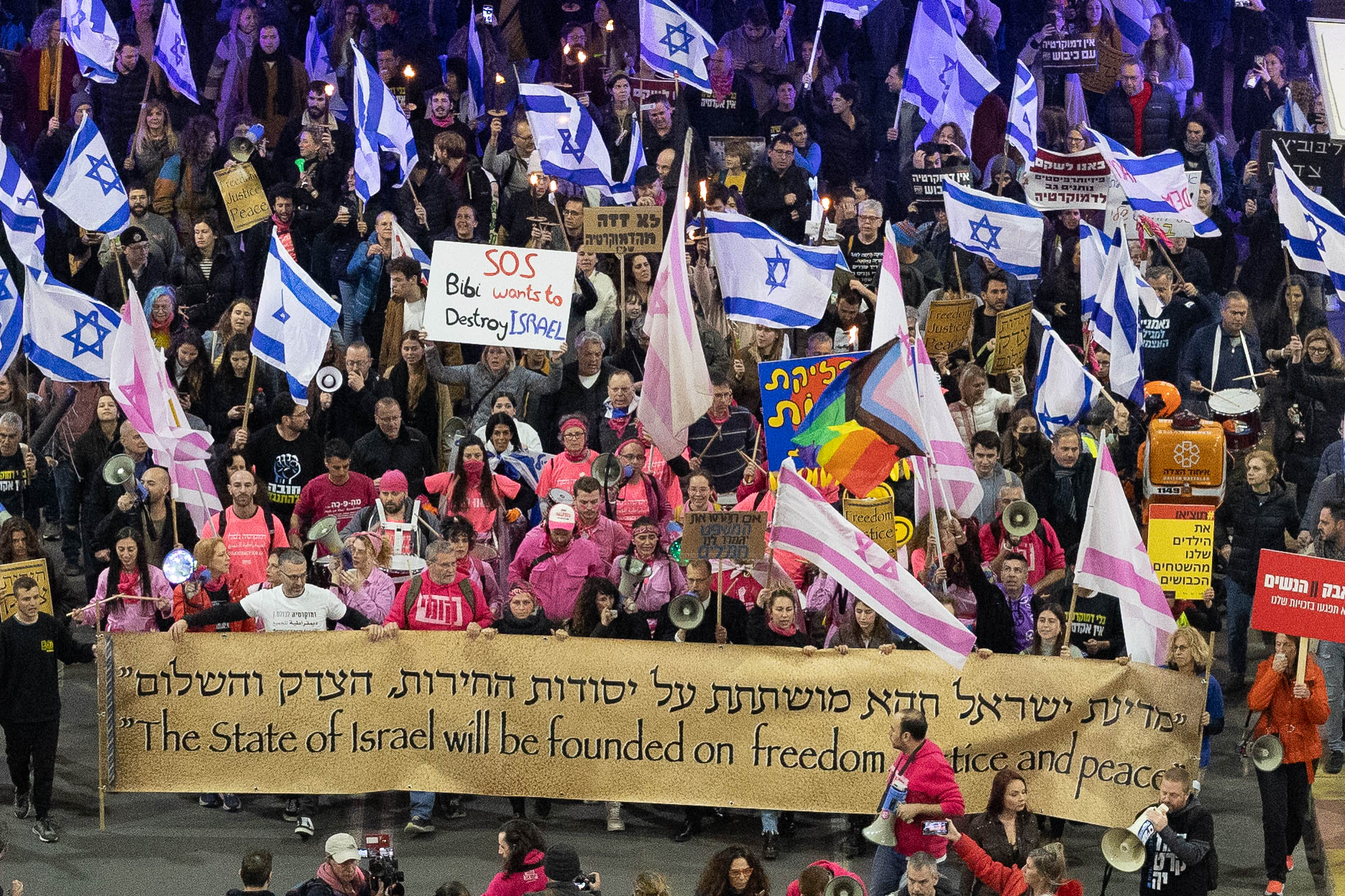 Photo by Erez Harodi of Israel democracy protest in 2023. Photo shows a large crowd of people holding Israeli flags. A large sign in the front reads "The state of Israel will be founded on freedom, justice, and peace" in both English and Hebrew. Another protest sign reads, "SOS Bibi wants to destroy Israel."