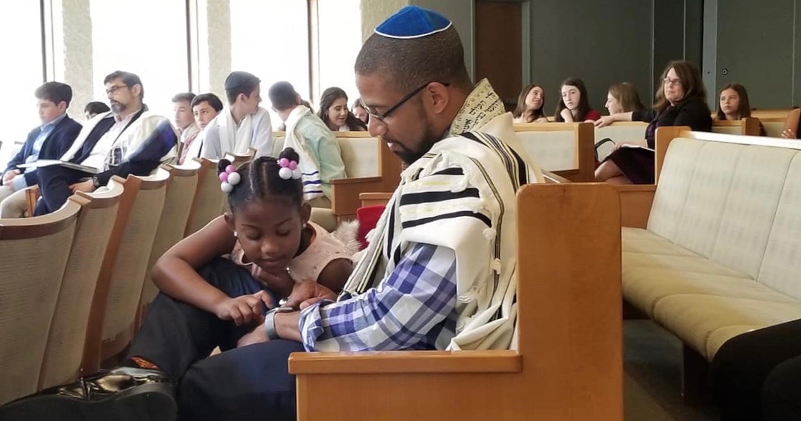 black male Jewish person wearing a tallit and blue kippah, sitting in the pews of a synagogue, as a little black girl in a pink dress sits on his lap. He is wearing glasses and looking down at her. Other congregants are in the background.