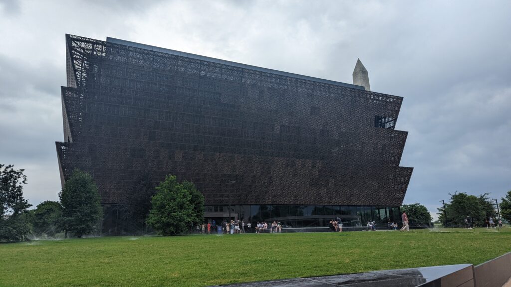 A modern, tiered museum building with a dark facade, trees, and people on the lawn; obelisk in background.