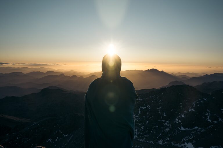 Person wrapped in a blanket watches the sun rise over mountains