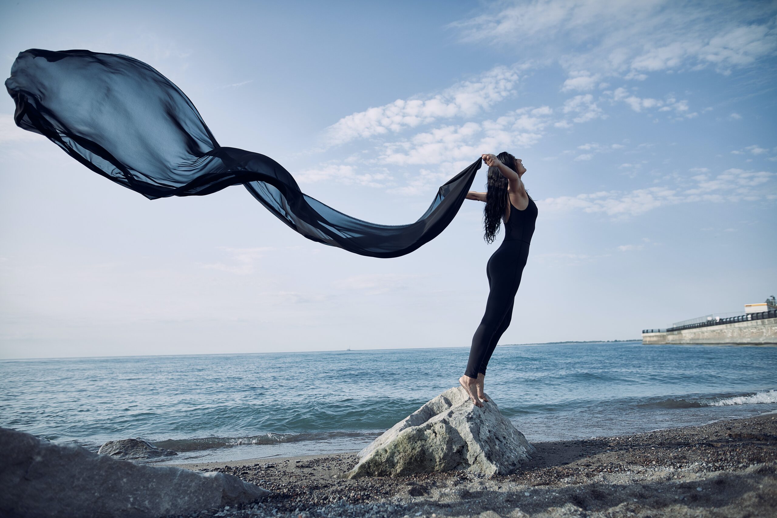 woman standing on a rock by the ocean with black scarf flowing behind her