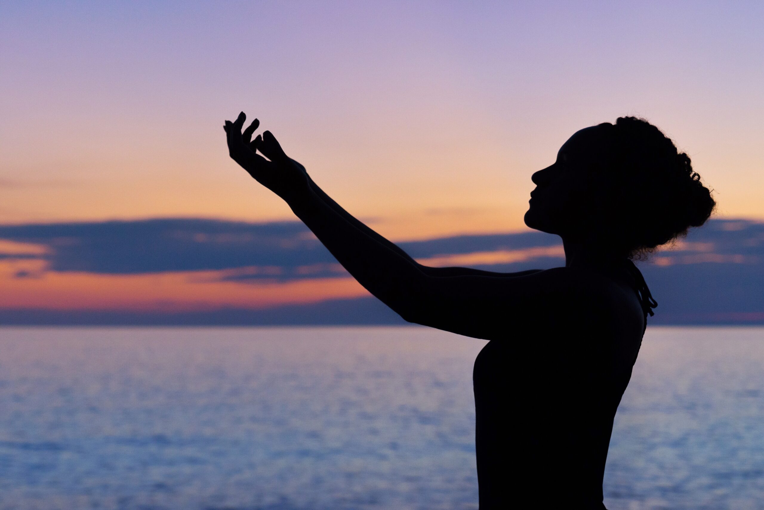 A silhouette of a person holding their arms against the sky, set against a body of water and sunset over mountains