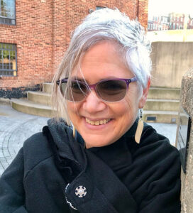 Smiling person with short gray hair and sunglasses, wearing a black coat, standing outside by brick buildings.