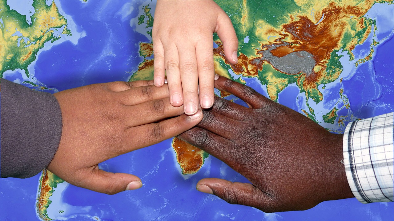 three hands--one with brown skin, one with white skin, one with black skin touch each other over the image of a world map