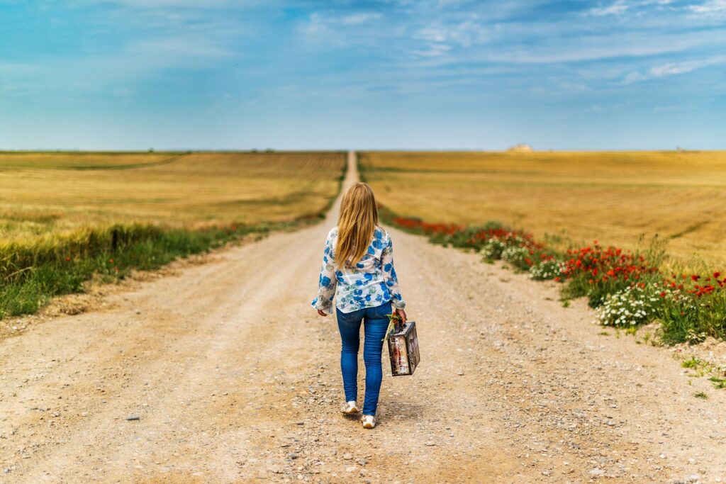 a girl holding a suitcase walks down a path surrounded by fields