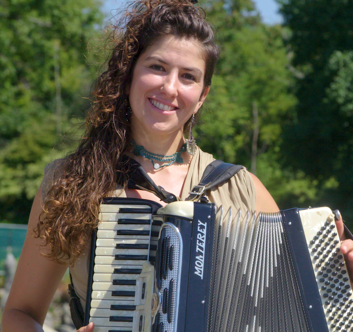 Eleanor has long curly brown hair. She holds an accordian and smiles at the camera