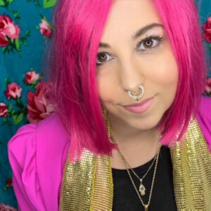 Anna Goodman Herrick is a white skinned woman with bright pink hair. She has a silver septum ring through her nose.