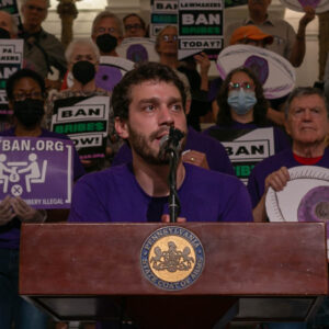 Miki Pollack is a white skinned man with brown haor, mustache and beard. he stands at a podium in fron tof a rally.