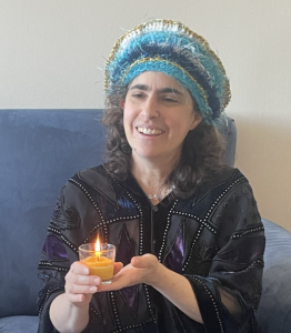 Rabbi Malkah Binah Klein is a white skinned woman wwearing a blue head covering. She holds a candle.