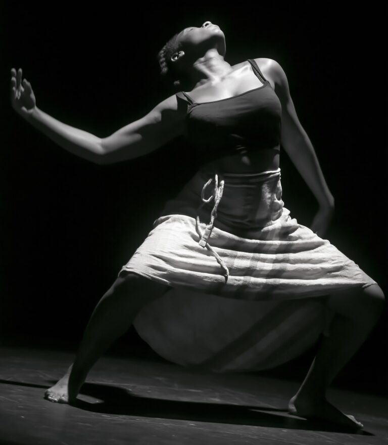 a black and white photo of a dancer with short hair wearing a long skirt with bent knees, face angled upward, and arm extending to the side