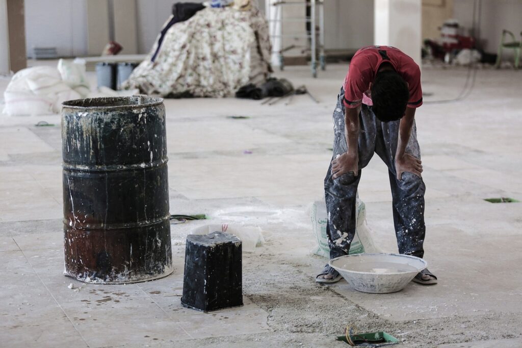 a construction scene in which a person wearing baggy jeans covered in paint and dust is hunched over a bowl, which is next to a bucket, a barrel, and a sack.