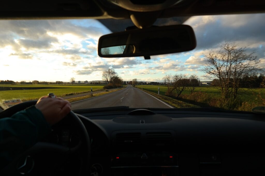 from inside the car we see a person's hands on a steering wheel and a rearview window, out of the window is pictured a road surrounded by grass and trees on a cloudy day