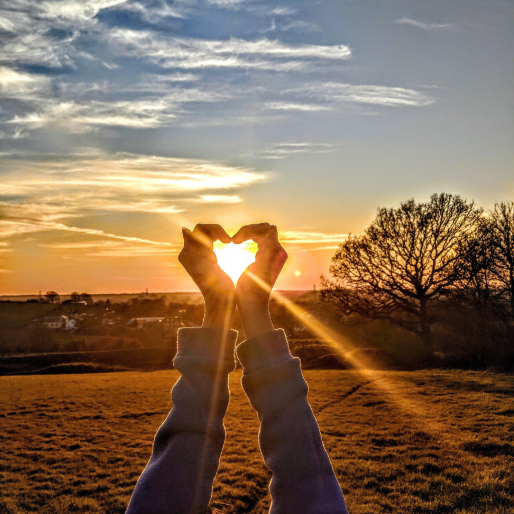 hands shaped as a heart, sunlight streaming through the heart. Set against a landscape of trees and blue sky with clouds and sun.