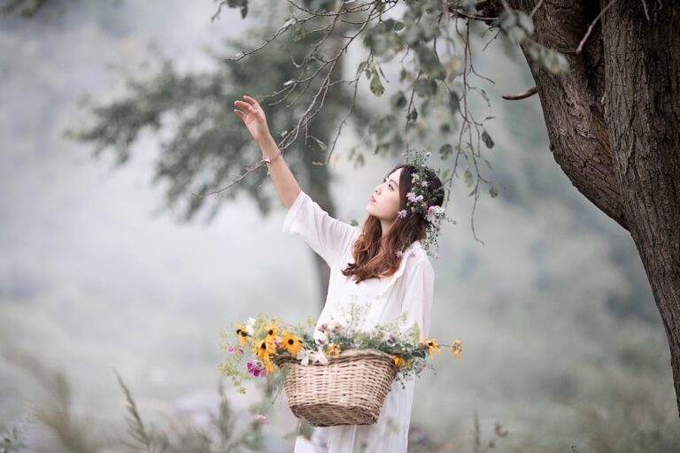 a woman in a white dress with a basket of flowers reaches up toward branches of a tree