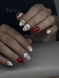 hands with painted nails: pinkies and pointer fingers are white with sparkly gold drip, ring fingers are all red, middle fingers have white background with apples that are cut open, thumbs have white backgrounds with red apples