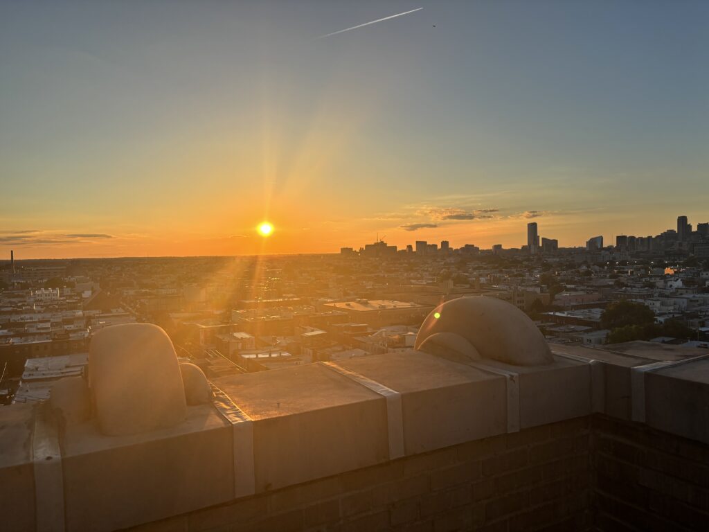 Bright orange sunset over a city skyline, viewed from a rooftop with stone ledges in the foreground.