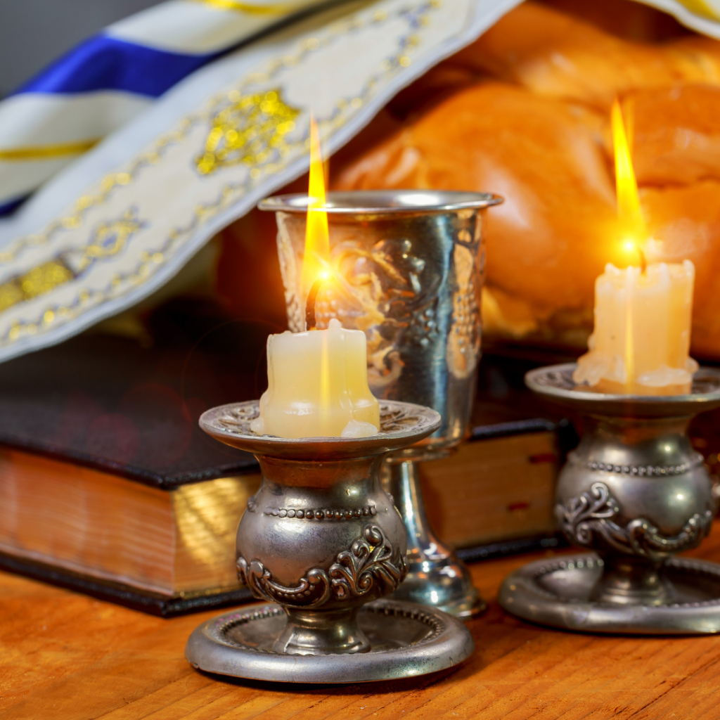 Two lit candles in ornate holders, a silver cup, a folded prayer shawl, and challah bread on a book.