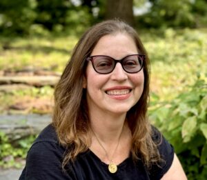 Gabrielle is a white-skinned womanwith long brown hair. She wars a black tshirt, sunglasses and a gold necklace. She sits in front of green trees and grass.