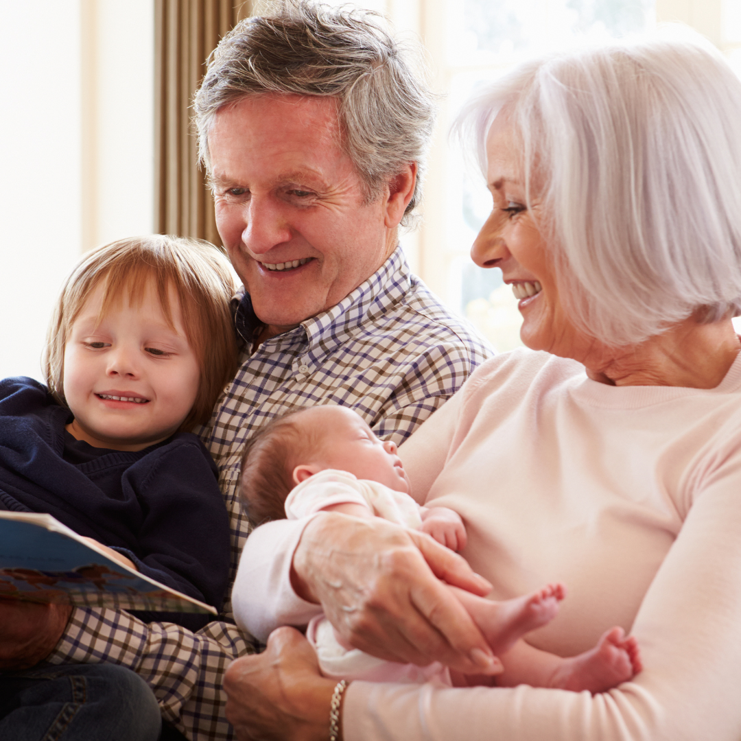 Two people with grey hair hold a toddler and an infant, reading them a book