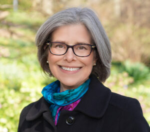 Smiling woman with glasses and gray hair, wearing a colorful scarf and black coat, outdoors with a blurred background.