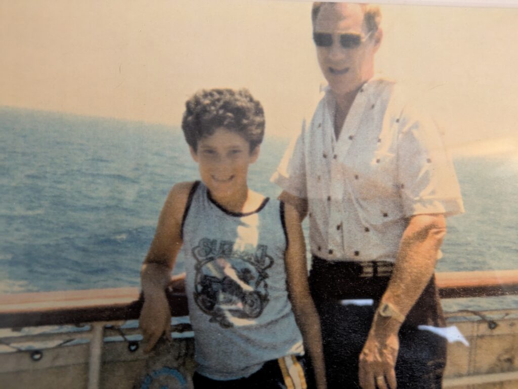 A boy and a man smiling on a boat with the ocean in the background.