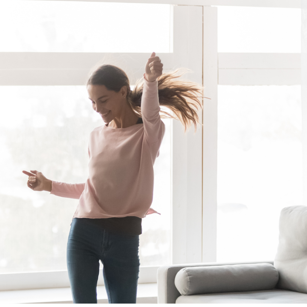 A joyful woman in a pink sweater and jeans dances in a bright living room with large windows. Her hair flows as she moves, and the room is softly lit with natural light.