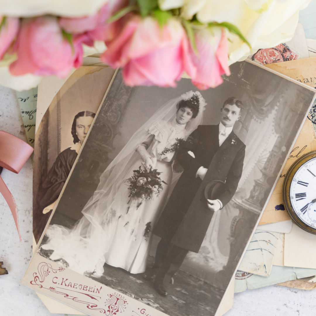 A vintage wedding photo of a bride and groom next to old letters, flowers, and a pocket watch.