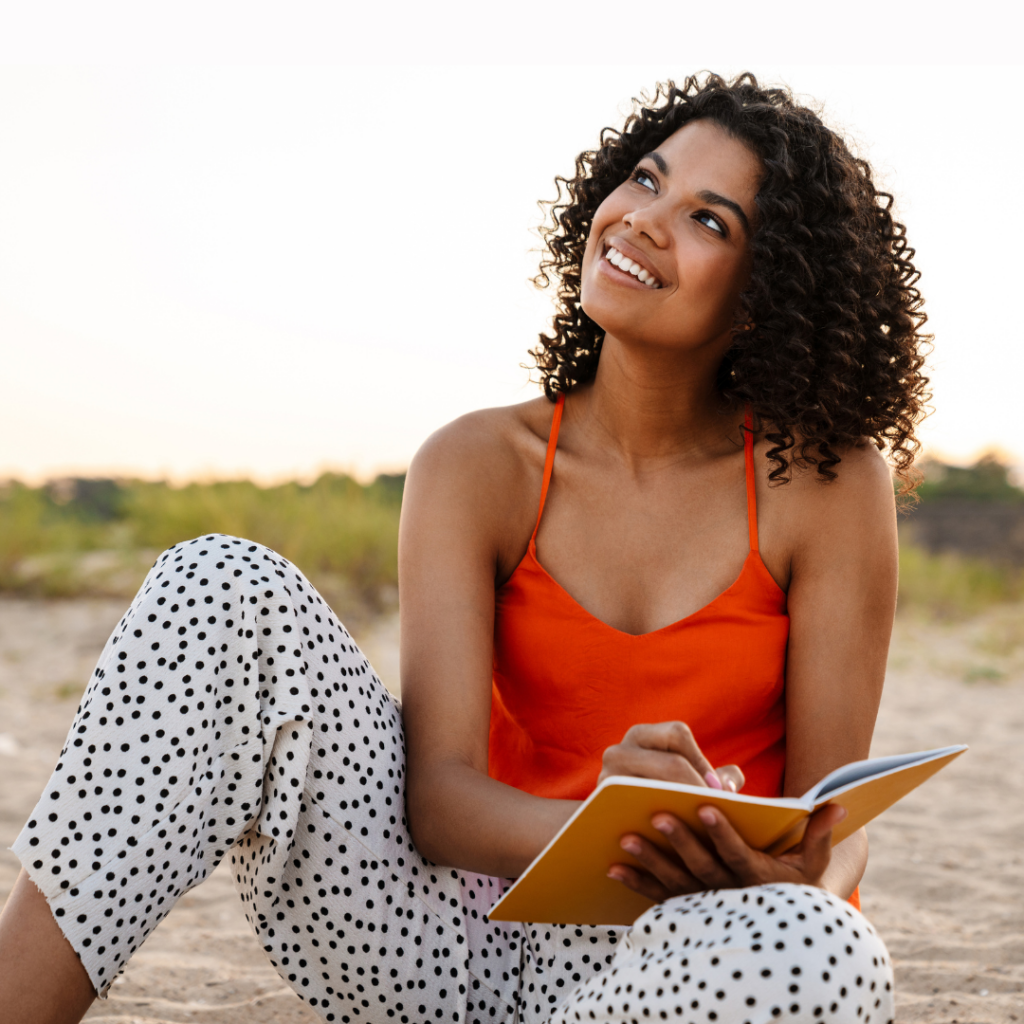 A woman in a red top and polka dot pants sits on a beach, smiling and writing in a notebook.