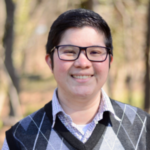 A smiling person with short hair, wearing glasses and a gray argyle sweater, stands outdoors with trees in the background.