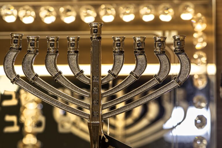 Close-up of a silver menorah with nine branches, set against a blurred, illuminated background.