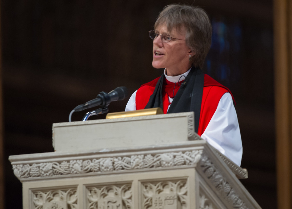 Rev. Budde in religious attire speaks at a podium with ornate carvings and a microphone.